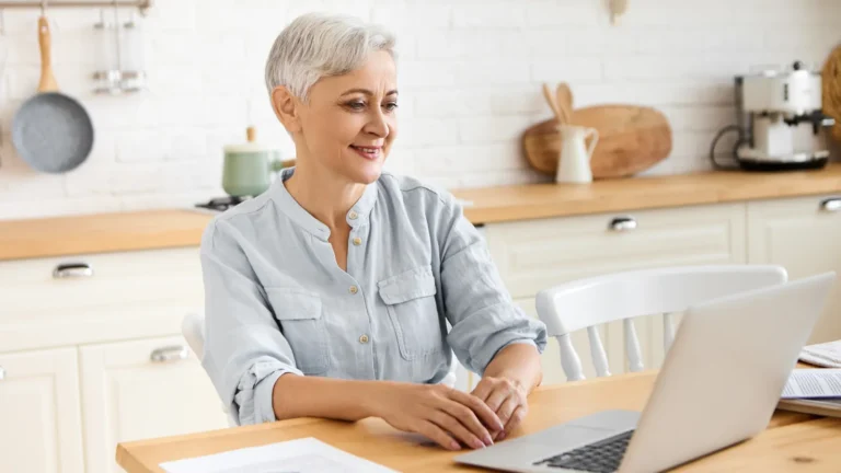 Older woman sitting at her kitchen table, smiling at her laptop